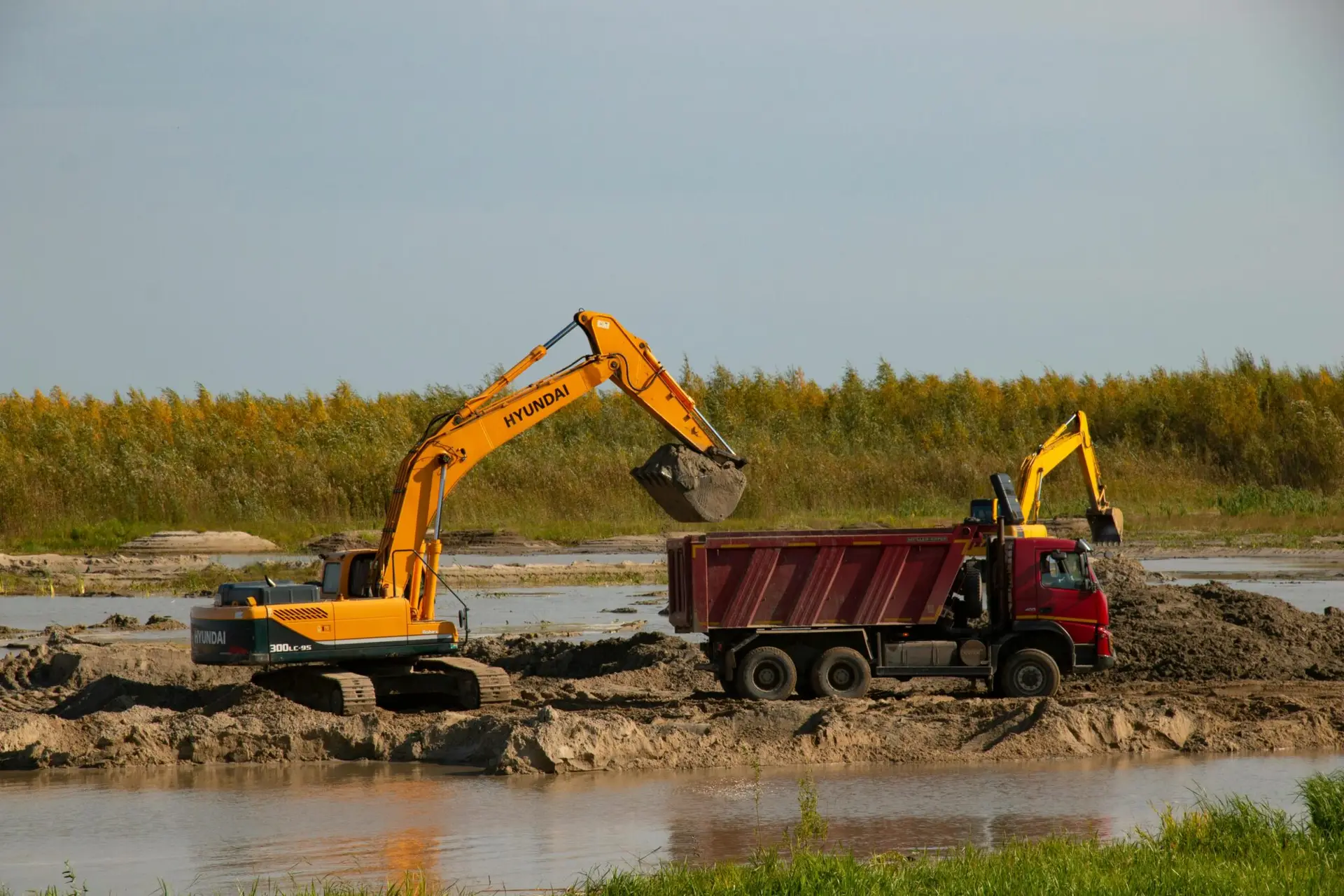 Excavator transfers soil to a dump truck by a river at a construction site. Machinery in action.
